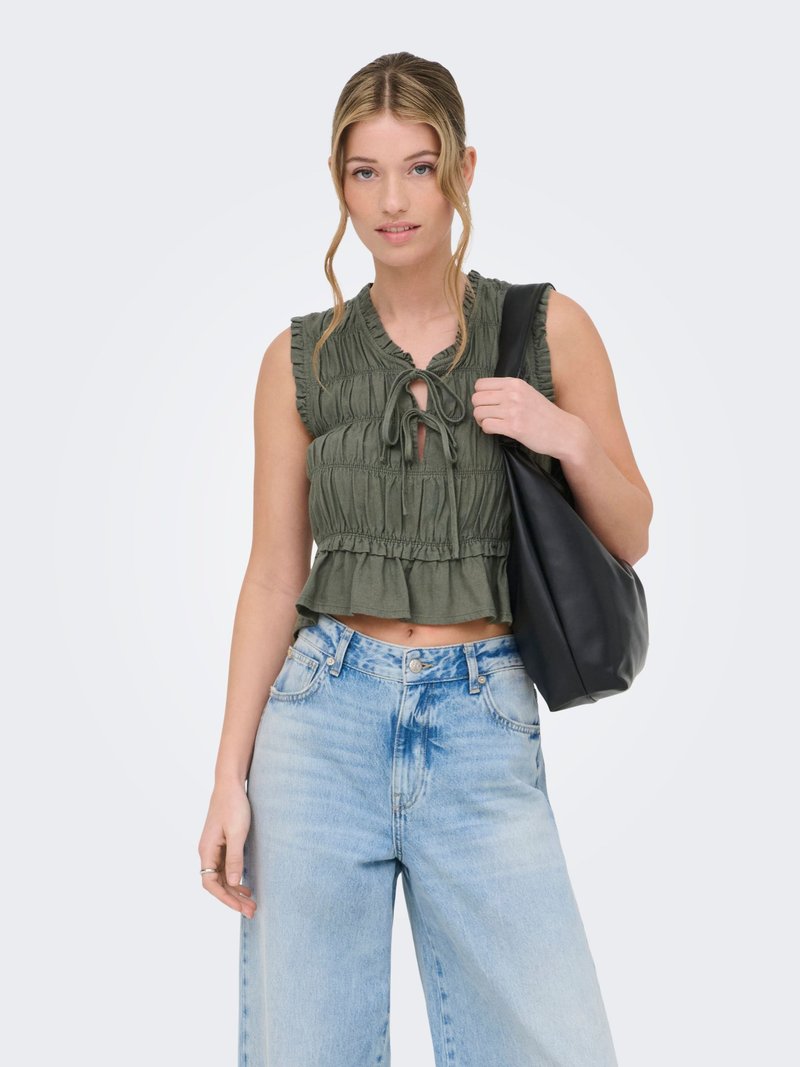 Giovane donna con capelli biondi sciolti, indossa un top verde oliva senza maniche con volant, jeans azzurri a vita alta e tiene una borsa nera a tracolla.