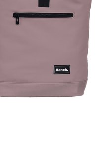 Sac à dos violet avec une finition mate élégante, présentant une poche zippée noire et une étiquette de logo noire. Accentuée par une poignée supérieure robuste.