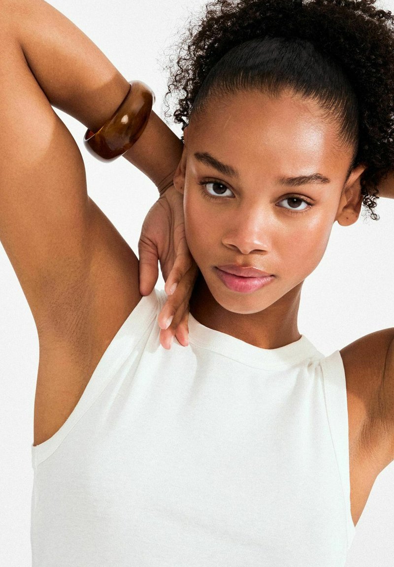 Bijeli top bez rukava s okruglim izrezom; koža na rukama vidljiva. Smeđi sjajni narukvica na jednom zapešću. Prirodne, teksturirane kovrče podignute visoko.