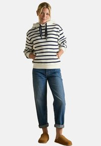 Kobieta stojąca z dłońmi w kieszeniach, ubrana w biało-granatowy, paskowany hoodie, podwinięte niebieskie dżinsy i brązowe buty wsuwane na tle białym.