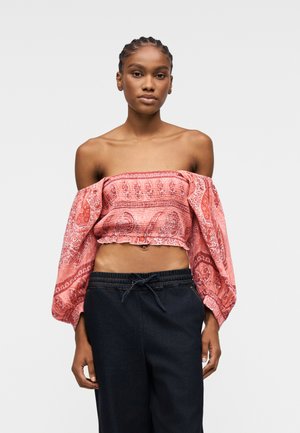 Jeune femme aux cheveux tressés portant un crop top rouge à motifs épaules dénudées et un pantalon sombre à cordon, debout devant un fond uni.