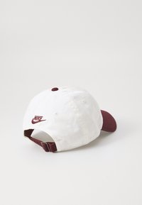 Bijela i bordo Nike bejzbol kapa s podešavanjem, izvezenim logom i šest rupa za ventilaciju na jednostavnoj svijetloj pozadini.