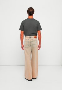 Homme aux cheveux bruns portant un t-shirt gris foncé et un jean beige à jambes larges, debout face à un mur blanc uni.