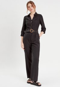 Jumpsuit di lino nero con colletto, chiusura a bottoni e maniche a tre quarti. Presenta una cintura intrecciata con una fibbia di legno rotonda.