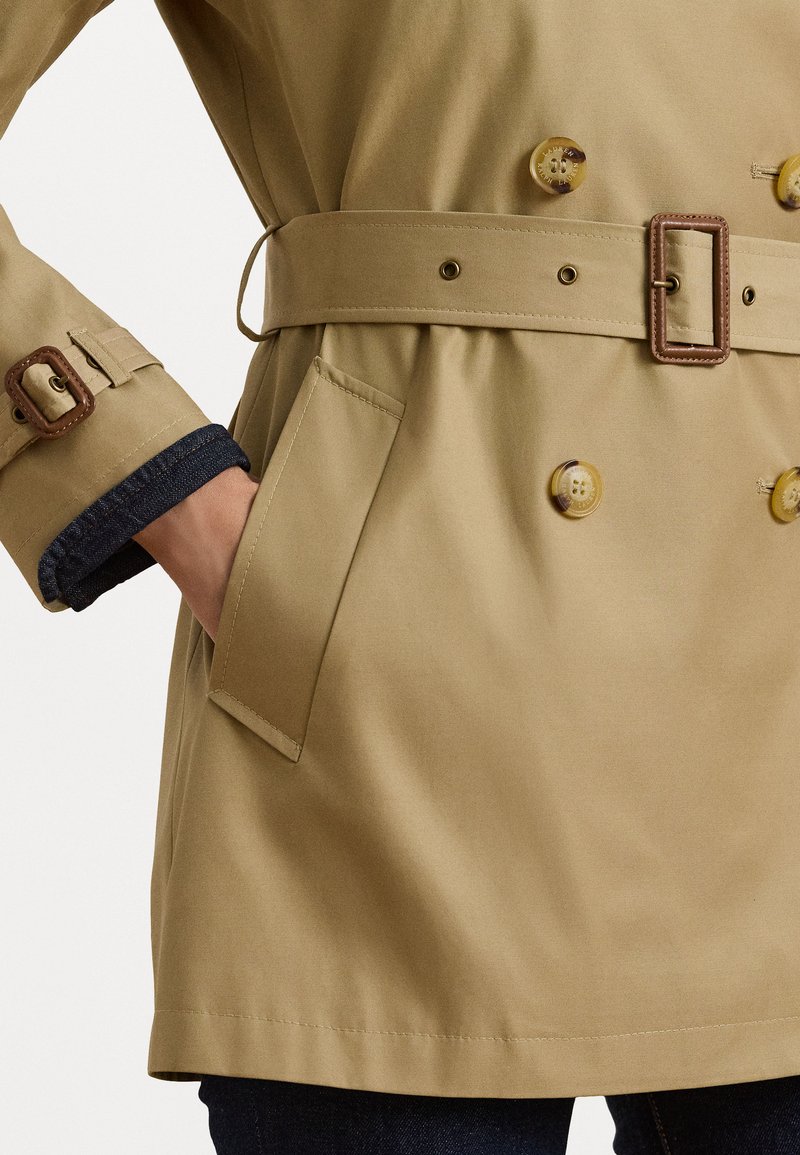 Beige trench coat with double-breasted design, matching belt, large buttons, and a side pocket. Features a textured fabric and cuffed sleeves.