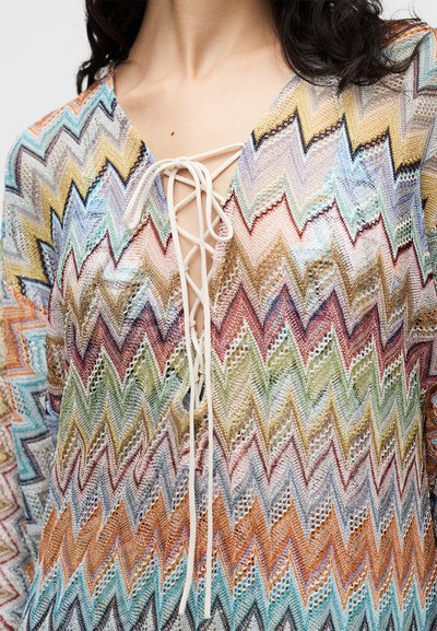 Gros plan d'un haut tricoté multicolore à motif zigzag avec un col en V lacé, porté par une personne aux cheveux foncés.