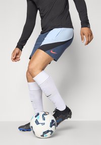 Man i svart skjorta och blå Nike-shorts som kontrollerar en vit och blå fotboll med svarta fotbollsskor och vita strumpor.