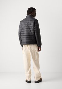 Zwarte, gewatteerde pufferjas met hoge kraag en gebreide manchetten. Gekleed over crèmekleurige broek met brede pijpen en donkere schoenen. Minimalistisch design.