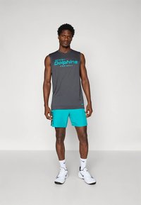 Grå ärmlös tröja med texten "Miami Dolphins Football", kombinerad med turkosa sportshorts. Vita sneakers och strumpor. Enfärgad bakgrund.