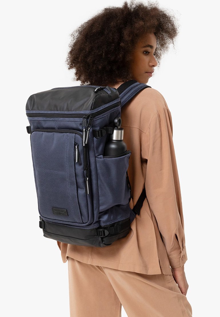 Sac à dos bleu marine en matériau texturé, doté d'un rabat supérieur, d'une poche latérale pour bouteille d'eau et de bretelles réglables. Accents de base noirs.
