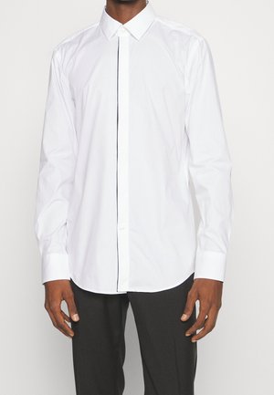 Mann trägt ein weißes Hemd mit langen Ärmeln und klassischem Kragen sowie schwarze Hose vor einem schlichten weißen Hintergrund.