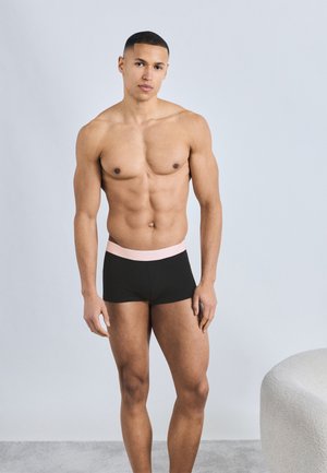 Αθλητικός άνδρας όρθιος, φοράει μαύρα κοντά εσώρουχα τύπου μπόξερ με ανοιχτό ροζ ζώνη μέσης, με φόντο ανοιχτό γκρι και μια καμπυλωτή καρέκλα κοντά.