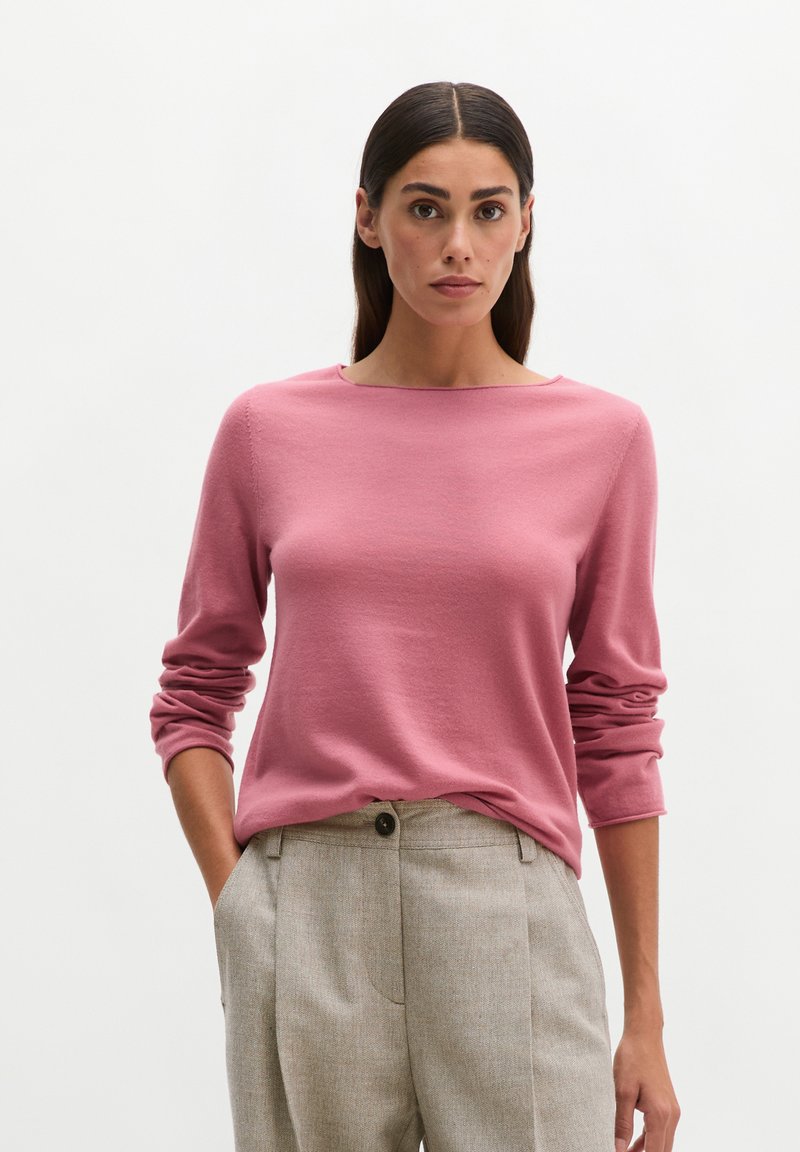 Woman with long dark hair wears a pink long-sleeve sweater and light beige high-waist trousers with hands in pockets against a plain background.