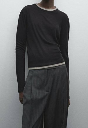 Personne portant un haut noir à manches longues avec bordure beige et pantalon large à rayures grises sur un fond clair uni.