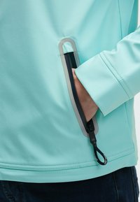 Giacca azzurro chiaro con una texture liscia, dotata di una tasca laterale con una rifinitura grigia e un tirante nero. Il polsino è aderente, consentendo libertà di movimento.