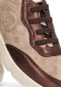 Zapatillas de ante beige con acentos metálicos marrones, logo redondo y cordones marrones. Superficie texturizada y diseño de suela redondeada y gruesa.