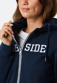 Hoodie zippé bleu marine en tissu doux, avec un lettrage "SIDE" brodé en blanc, une fermeture éclair à l'avant et une capuche réglable avec des cordons.