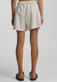 Shorts en lin couleur crème avec une taille élastique et une coupe décontractée. L'ourlet est coupé à cru, offrant un look informel. Portés avec des sandales marron.