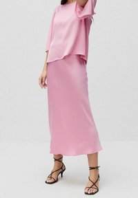 Nainen, jolla on yllään rento vaaleanpunainen satiinipusero, joka on tippunut yhteen saman sävyisen pitkän satiinihameen kanssa, ja mustat nyörisaappaat, seisoo valkoisella lattialla.