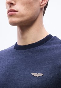 Νεαρός ενήλικας που φοράει μπλε σκούρο υφασμάτινο πουκάμισο με μικρό λογότυπο Aston Martin σε μπεζ χρώμα στο στήθος, εικονιζόμενος από τον λαιμό μέχρι τον πάνω κορμό.