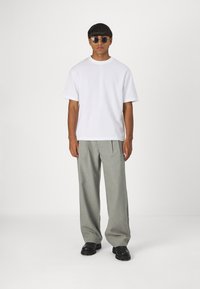 Weißes T-Shirt mit kurzen Ärmeln, graue weite Hosen und schwarze Schuhe. Model trägt runde Sonnenbrillen und steht vor einem schlichten Hintergrund.