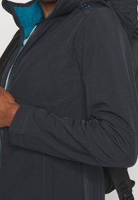 Chaqueta azul marino oscuro con una textura suave, capucha y forro en turquesa contrastante. Presenta un zipper frontal y se lleva con una mochila.