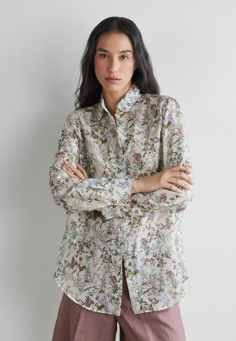 Vrouw met lang donker haar, gekleed in een bloemenblouse met knopen en mauve broek, staat met gekruiste armen tegen een effen witte achtergrond.