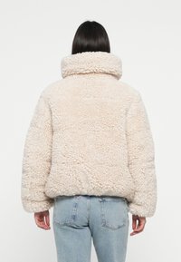 Fuzzy beige shearling jasje met een hoge kraag, oversized vorm en een gestructureerd oppervlak. Gecombineerd met lichtblauwe denim jeans.