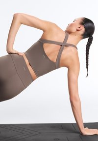 Žena s pletenými vlasmi vykonáva bočný plank na čiernej joga podložke, má na sebe hnedý športový podprsenku s krížovými popruhmi a zodpovedajúce legíny.