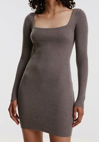 Kvinna klädd i en figursydd, ribbad, långärmad klänning i taupe med fyrkantig halsringning mot en enfärgad vit bakgrund.