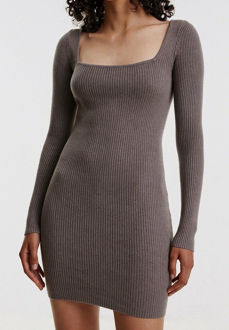 Kvinna klädd i en figursydd, ribbad, långärmad klänning i taupe med fyrkantig halsringning mot en enfärgad vit bakgrund.