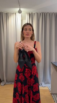 Γυναίκα με μαύρο φόρεμα με κόκκινα τριαντάφυλλα κρατάει μαύρα μυτερά παπούτσια, στέκεται σε εσωτερικό χώρο με φωτεινές κουρτίνες και ξύλινο πάτωμα ορατό πίσω της.