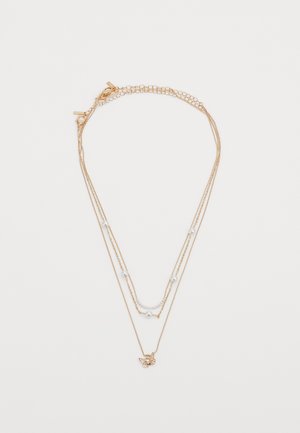 Aranyszínű, réteges nyaklánc, amely három szálból áll: két szálon kis fehér gyöngyök, egy szálon pedig egy pillangó medál található, bonyolult lánc kialakítással.