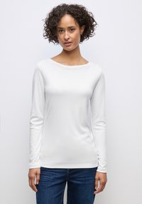 Weißes Langarmhemd mit weitem Ausschnitt und glatter Textur, kombiniert mit dunkelblauen Jeans. Einfaches, tailliertes Design.