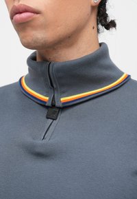 Pele de velo cinzenta com fecho de correr, com gola alta, apresentando um detalhe de riscas multicoloridas e um puxador de fecho preto. Textura suave com um ajuste justo.