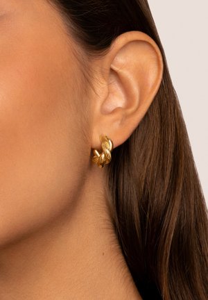 Gouden oorbellen met een gedraaide ontwerpen, met gladde en gestructureerde oppervlakken, bevestigd aan het oor. Close-up van het sieraad.
