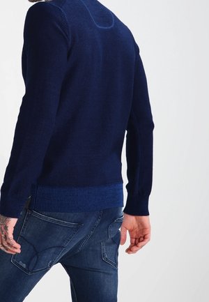 Dunkelblauer gestrickter Pullover mit geripptem Saum und Bündchen. Verfügt über einen runden Halsausschnitt und strukturierte Details auf der Rückseite. Kombiniert mit blauen Jeans.