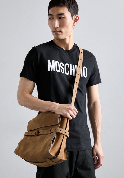 Geantă de umăr din suede maro, cu clapetă, detaliu cu cataramă și curea ajustabilă. Modelul poartă un tricou negru cu logo-ul Moschino.