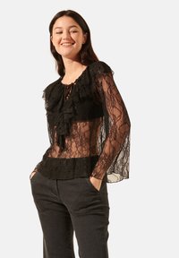 Blouse en dentelle noire avec des accents froncés, des manches longues transparentes et un col à nouer, associée à un pantalon noir dans une posture décontractée.