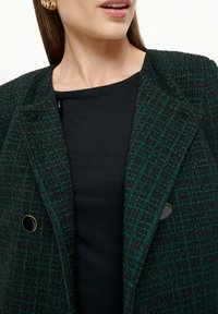 Zeleni i crni karirani tweed sako s dva crna gumba, nosi se preko crne majice, a karakterizira ga teksturirana tkanina i strukturirana ramena.