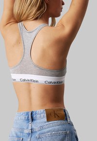 Brassière gris con un diseño deportivo y una banda blanca con el texto "Calvin Klein", combinada con pantalones cortos de mezclilla azul y una etiqueta de cuero.