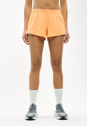 Femeie purtând pantaloni scurți atletici portocalii Under Armour, șosete albe până la gleznă și pantofi gri de alergare, stând pe un fundal alb.