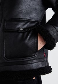 Black faux leather jacket featuring a textured finish, fleece-lined collar and cuffs, and a buttoned chest pocket. Red logo accent on the side.