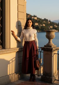 Biały, dziergany sweter z golfem w połączeniu z bordową plisowaną spódnicą, brązową skórzaną torebką i kozakami za kolano; elegancka sceneria na zewnątrz.