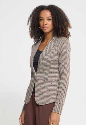 Jeune femme aux cheveux bouclés porte un blazer beige à carreaux sur un haut noir et un pantalon marron, debout devant un fond uni.