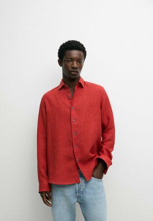 Ein Mann trägt ein rotes Hemd mit Knöpfen und hellblaue Jeans und steht vor einem schlichten weißen Hintergrund mit einer Hand in der Hosentasche.