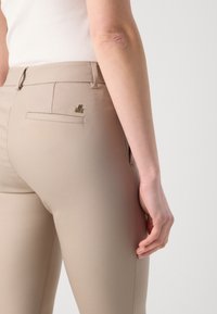 Personne portant un pantalon beige ajusté et un haut blanc sans manches, montrant une cicatrice sur l'arrière du bras droit près du coude.