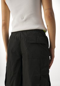 Vue arrière d'une personne portant un pantalon cargo noir avec de grandes poches et un haut blanc sans manches à côtes. Les tatouages indiquent « Berlin » et « São Paulo ».