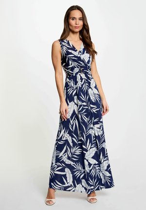 Mujer de pie con un vestido maxi sin mangas azul marino con estampado de hojas tropicales blancas y cinturón en la cintura, combinado con tacones blancos abiertos en la punta.