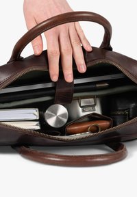 Brown leather bag with a zipper, containing notebooks, a metal water bottle, a wallet, and a laptop, with a hand interacting with it.